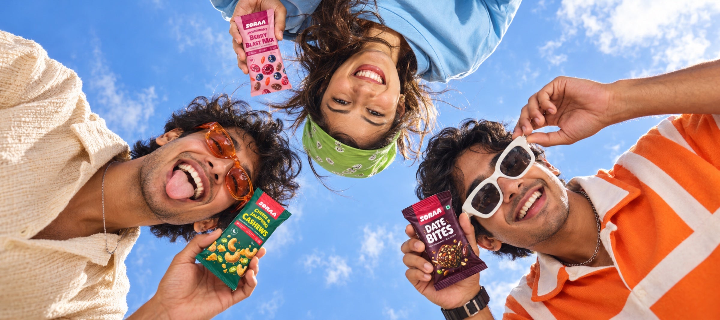 Three friends enjoying SORAA Berry Blast Mix, Cheese and Jalapeno Cashews, and Date Bites beneath a blue sky